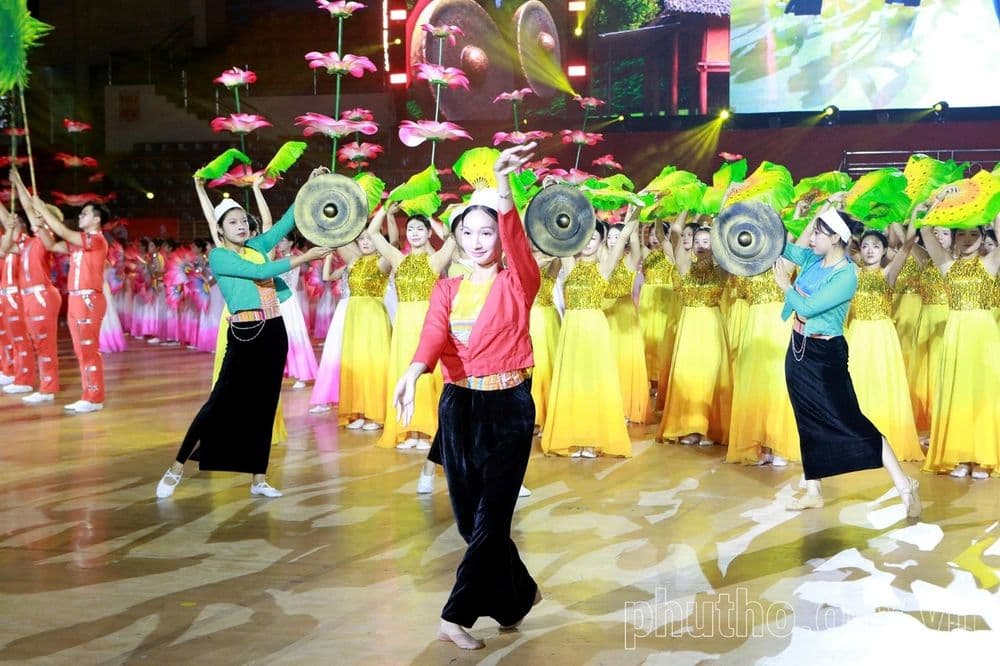 Vũ công trong trang phục dân tộc cầm chiêng múa ở tiền cảnh, phía sau là khối diễn viên áo vàng và đạo cụ hoa sen dưới ánh đèn sân khấu