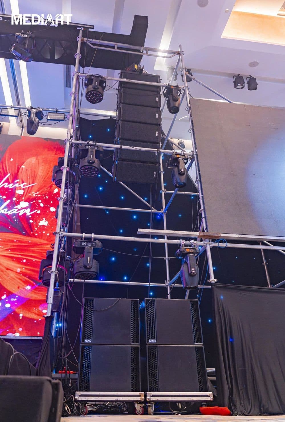 Toàn cảnh hệ thống NEXO của BOS Entertainment: dàn line array GEO S12 treo trên tháp giàn giáo, hai cụm sub LS18 xếp chồng bên dưới, phông sao và đèn moving head tại đêm nhạc trong nhà
